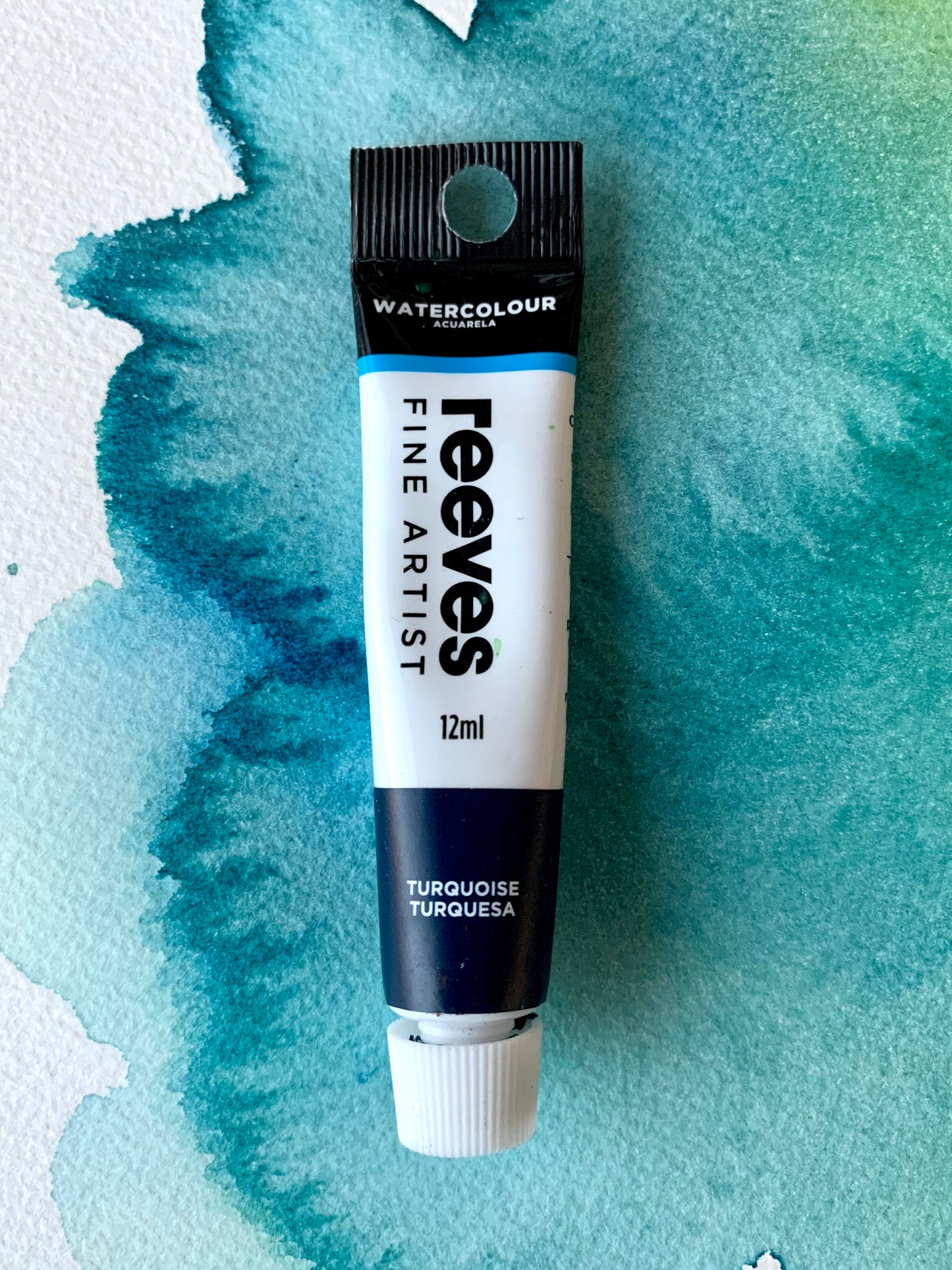 A 12ml tube of Reeves Fine Artist Watercolour paint in the shade Turquoise, placed on a textured watercolor paper background with blue and green washes.