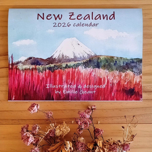 2026 New Zealand calendar with a mountain and red flower field illustration on a wooden surface.